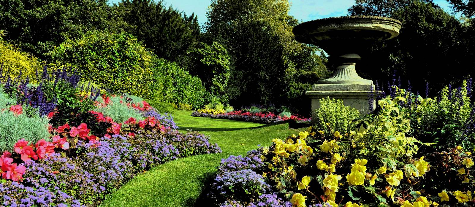 A lush garden with manicured green lawns and vibrant flowerbeds in red, purple, yellow, and blue showcases expert backyard design. A large stone urn sits prominently to the right, surrounded by blooming plants. Trees form a dense backdrop under a partly cloudy sky.