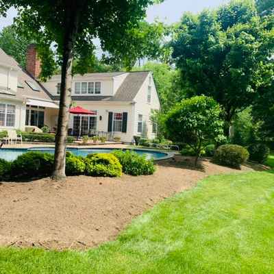 Backyard view of a two-story house with large windows. A circular swimming pool, surrounded by neatly landscaped shrubs and trees, showcases exquisite hardscape design. Patio furniture and a red umbrella near the pool highlight expert property maintenance, with well-maintained grass under a clear sky.