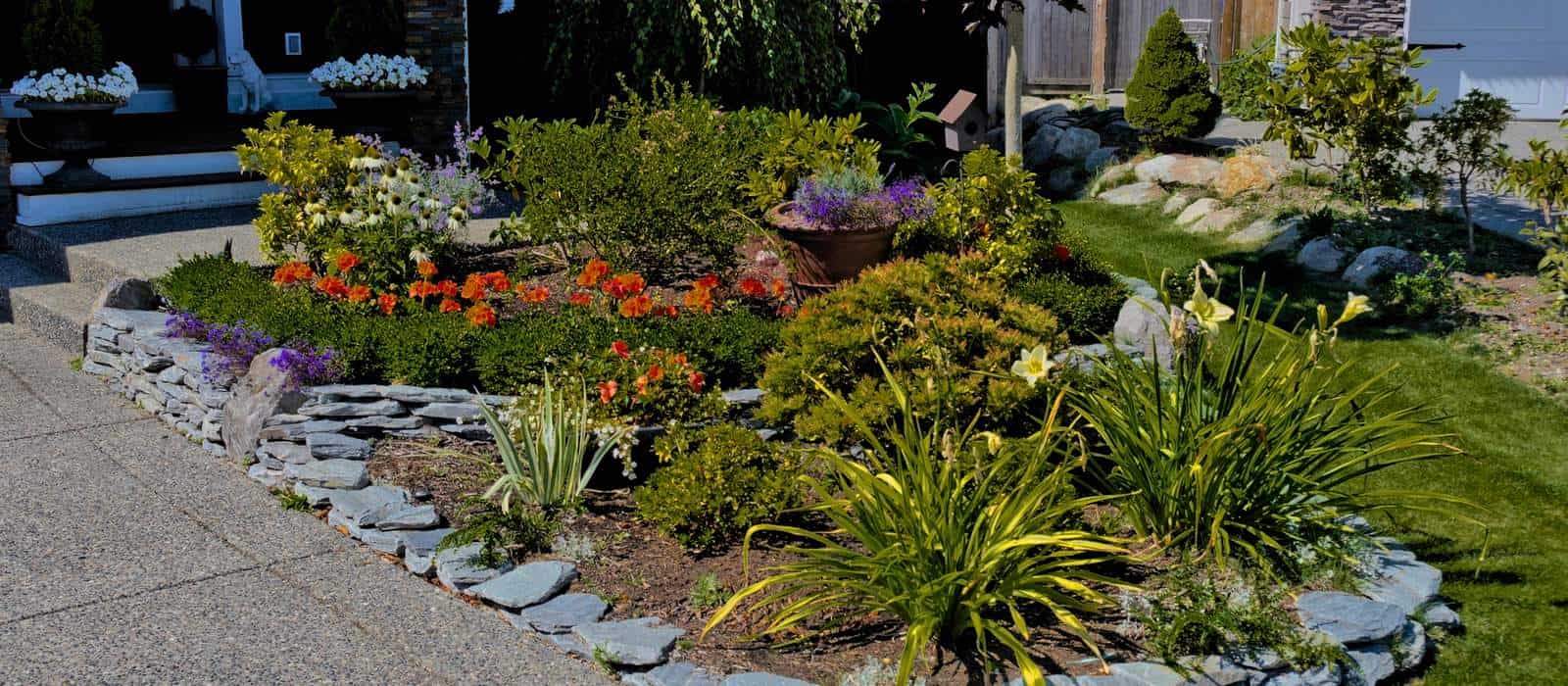 A neatly landscaped garden showcases a variety of colorful flowers, including orange and purple blooms, surrounded by a low stone border. Green shrubs and plants complement the backyard design, while a paved walkway is visible in the foreground, reflecting meticulous yard maintenance.