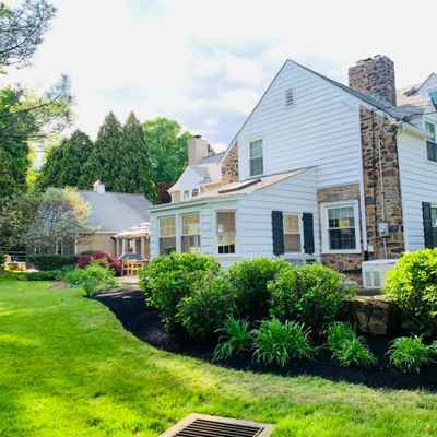 A scenic view of a white house with stone accents, surrounded by lush greenery showcases impeccable yard maintenance. The elegant hardscape design complements gardens, bushes, and a grassy lawn. Tall trees in the background frame the bright, sunny day beautifully.