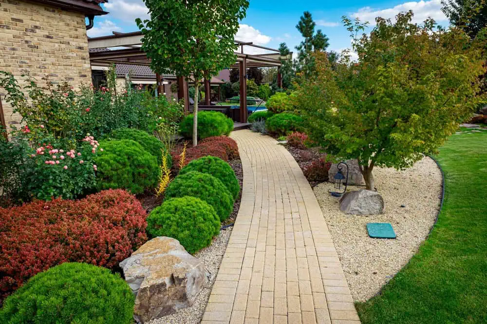 A garden path made of beige bricks winds through a landscaped yard, crafted by skilled landscapers in Delaware County. Lush green and red shrubs, flowering plants, and small trees border the walkway. A brick house is visible in the background under a partly cloudy sky.