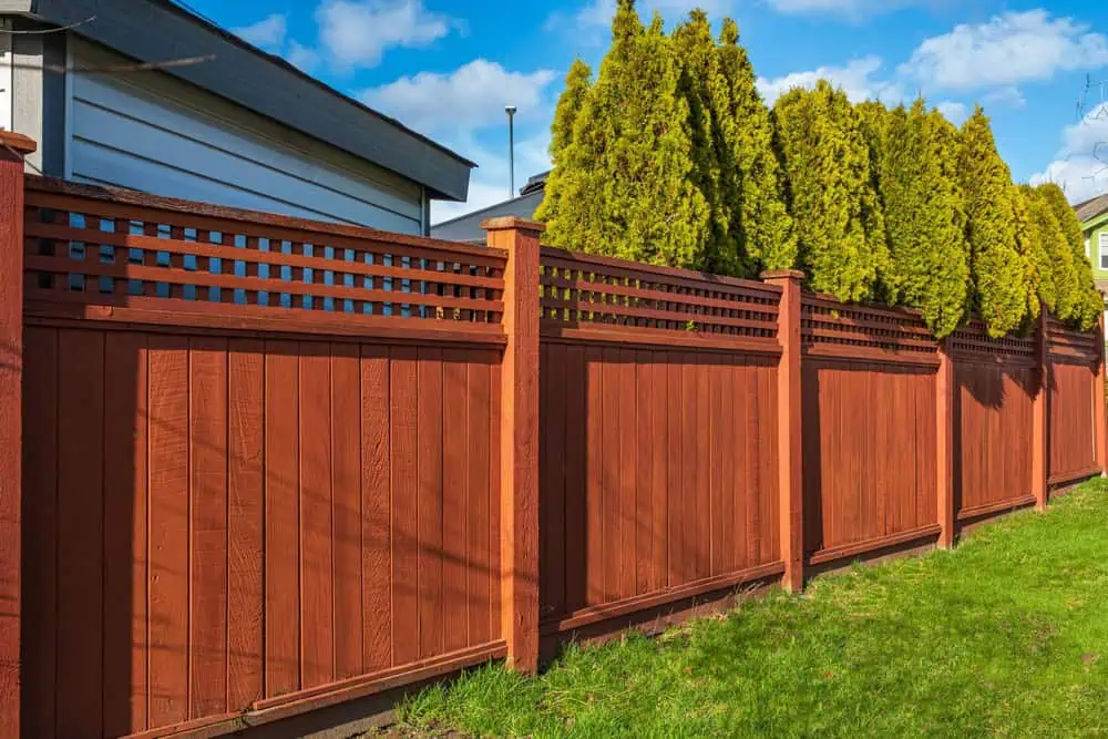 A tall, wooden privacy fence with decorative lattice tops, painted in a rich brown-red color, runs alongside a green grassy yard. Thanks to expert fence installation in Delaware County, evergreen trees and part of a house are visible under a blue sky with clouds.