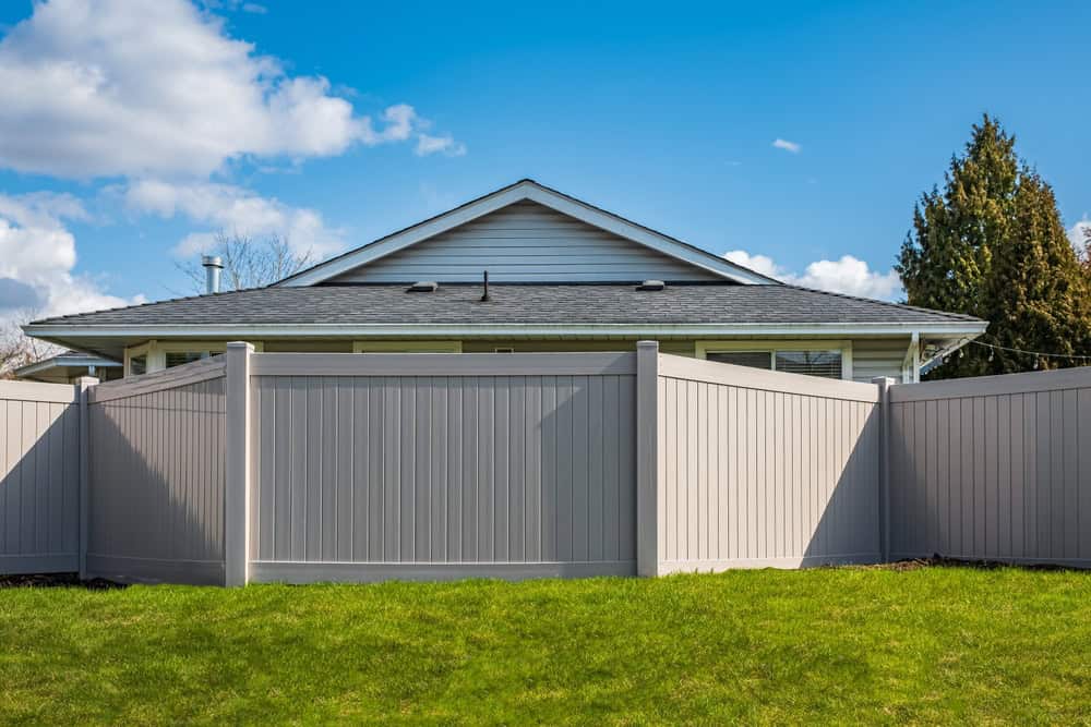 A house with a gray roof is partially visible behind a tall, beige privacy fence. Expertly crafted through fence installation in Delaware County, the structure encloses a green lawn under a clear blue sky dotted with fluffy clouds.