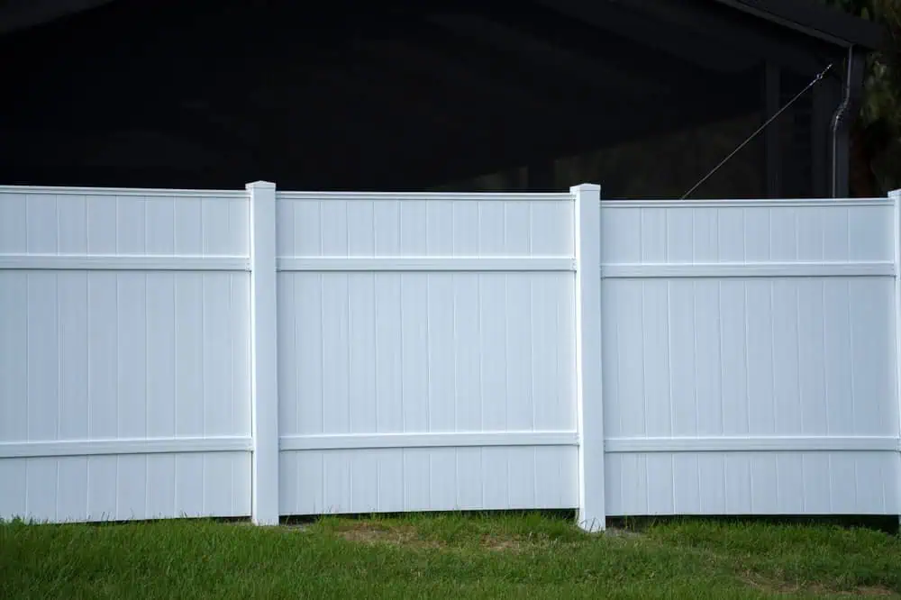 A white vinyl privacy fence with three vertical panels stands on a grassy lawn. Behind the fence, part of a dark structure is visible. The scene is brightly lit, highlighting the clean lines and simple design of the fence.