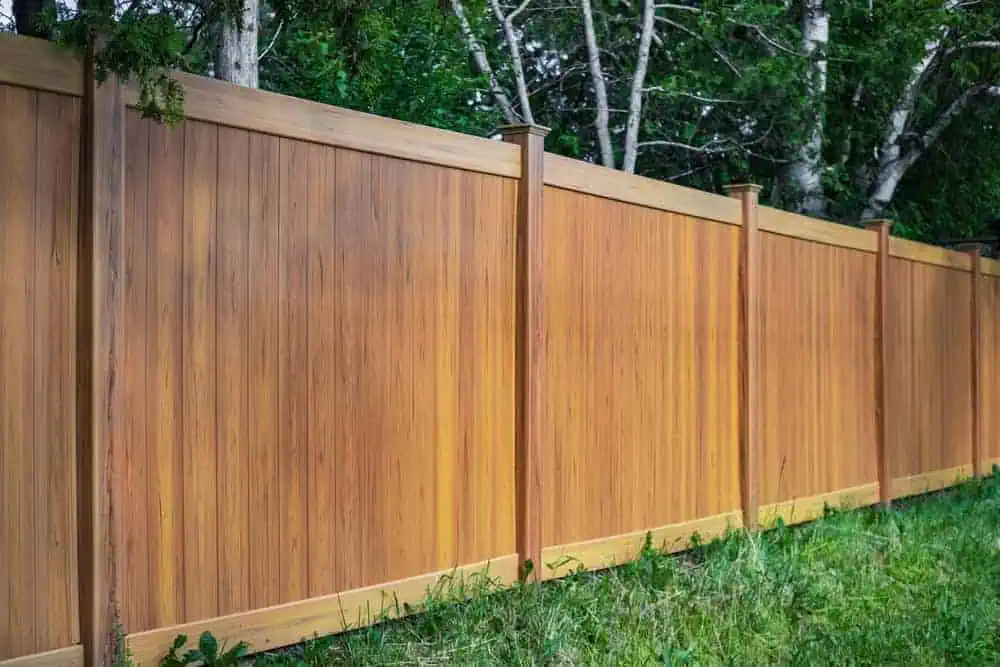 A long, wooden privacy fence runs along a grassy area, bordered by dense green trees. The fence panels are a warm brown color, supported by evenly spaced vertical posts, creating a natural and serene outdoor setting.