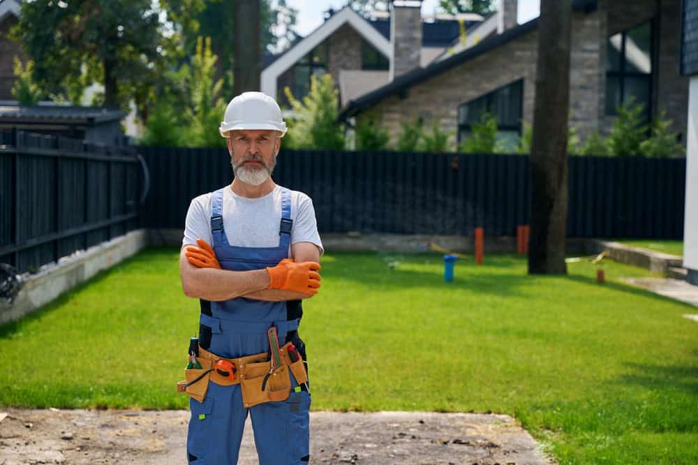 A construction worker with a white beard stands confidently in a backyard, wearing a white hard hat, orange gloves, and blue overalls with a tool belt. The background features a modern house and a green lawn.