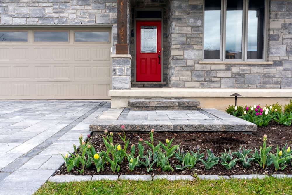 Front view of a house with gray stone exterior, beige garage door, a red front door, stone steps, and a flower bed with budding tulips along the entrance path.