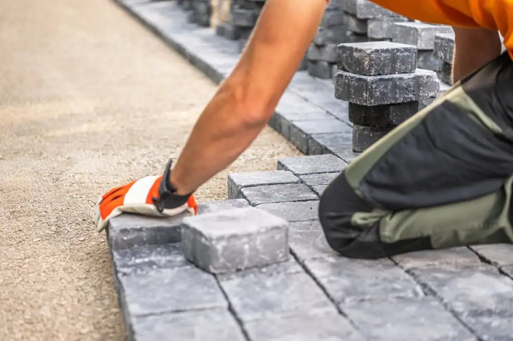 A person wearing an orange shirt and work gloves is kneeling and laying gray paving stones in a pattern on a sandy surface. Several stacks of stones are visible nearby, indicating ongoing construction.