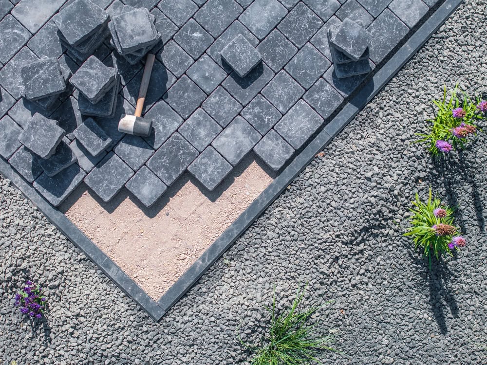 A section of a patio being constructed with dark paving stones. A rubber mallet rests on the partially laid tiles. The surrounding area is covered with gravel and features small patches of greenery with purple flowers.