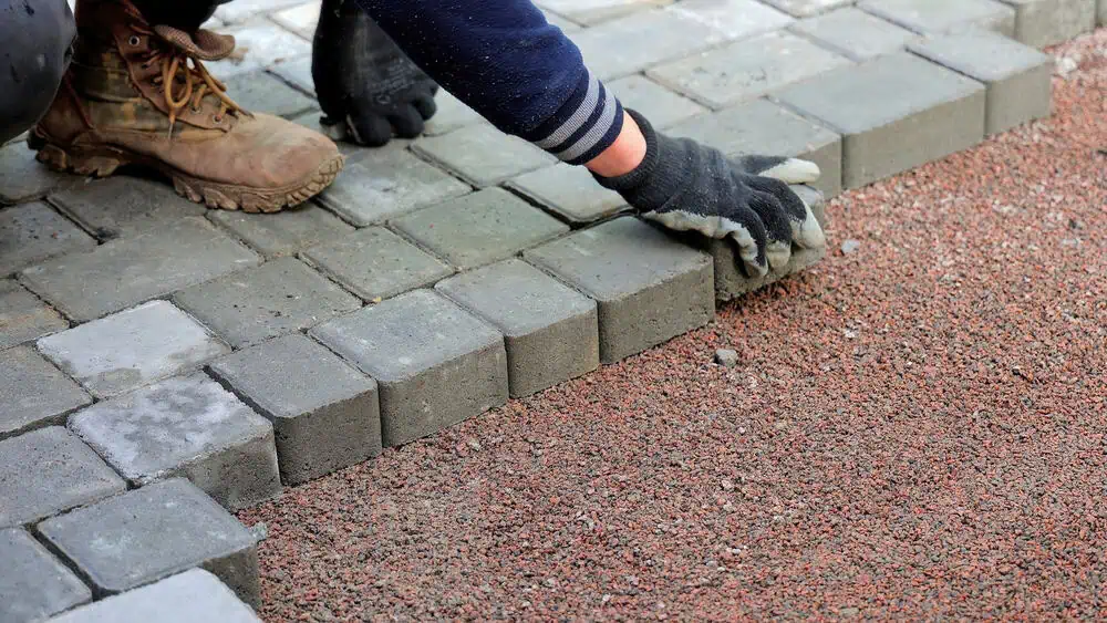 A person wearing gloves and work boots is laying gray paving stones on a sandy surface. The person's sleeve is visible as they adjust the stones, creating a neat pattern. The surrounding area has unpaved brown gravel.